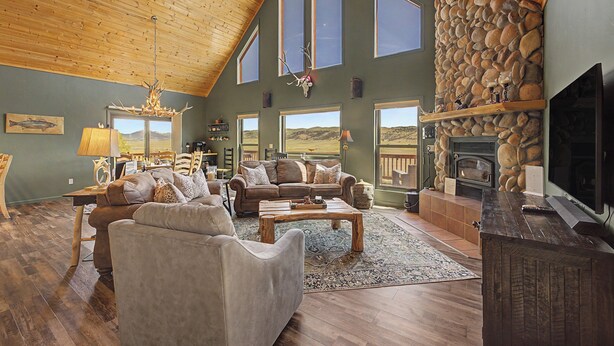 A spacious living room with a high ceiling, stone fireplace, large windows, and a television. - Trout Fishing & Panoramic Views - Starlink, 4BR, Pet-Friendly (Fairplay)