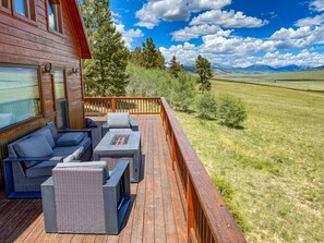 A wooden deck with outdoor furniture, including a sofa and chairs, overlooking a grassy landscape with trees and mountains.