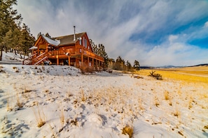 A log cabin with a porch and a chimney, surrounded by snow and a field.