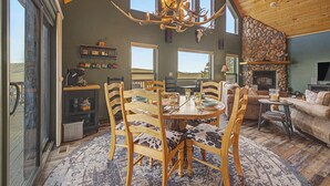 A spacious living room with a large dining table, wooden chairs, a fireplace, and a stone wall. - Trout Fishing & Panoramic Views - Starlink, 4BR, Pet-Friendly (Fairplay)