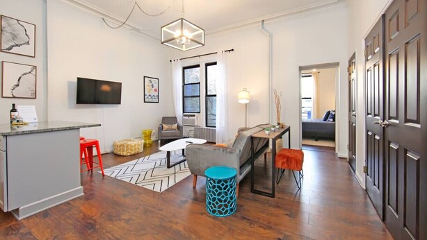 Una sala de estar moderna con un sofá gris, una mesa de centro y un televisor. Se observa una zona de cocina con una encimera gris y un taburete rojo. El cuarto tiene piso de madera y una alfombra estampada.
