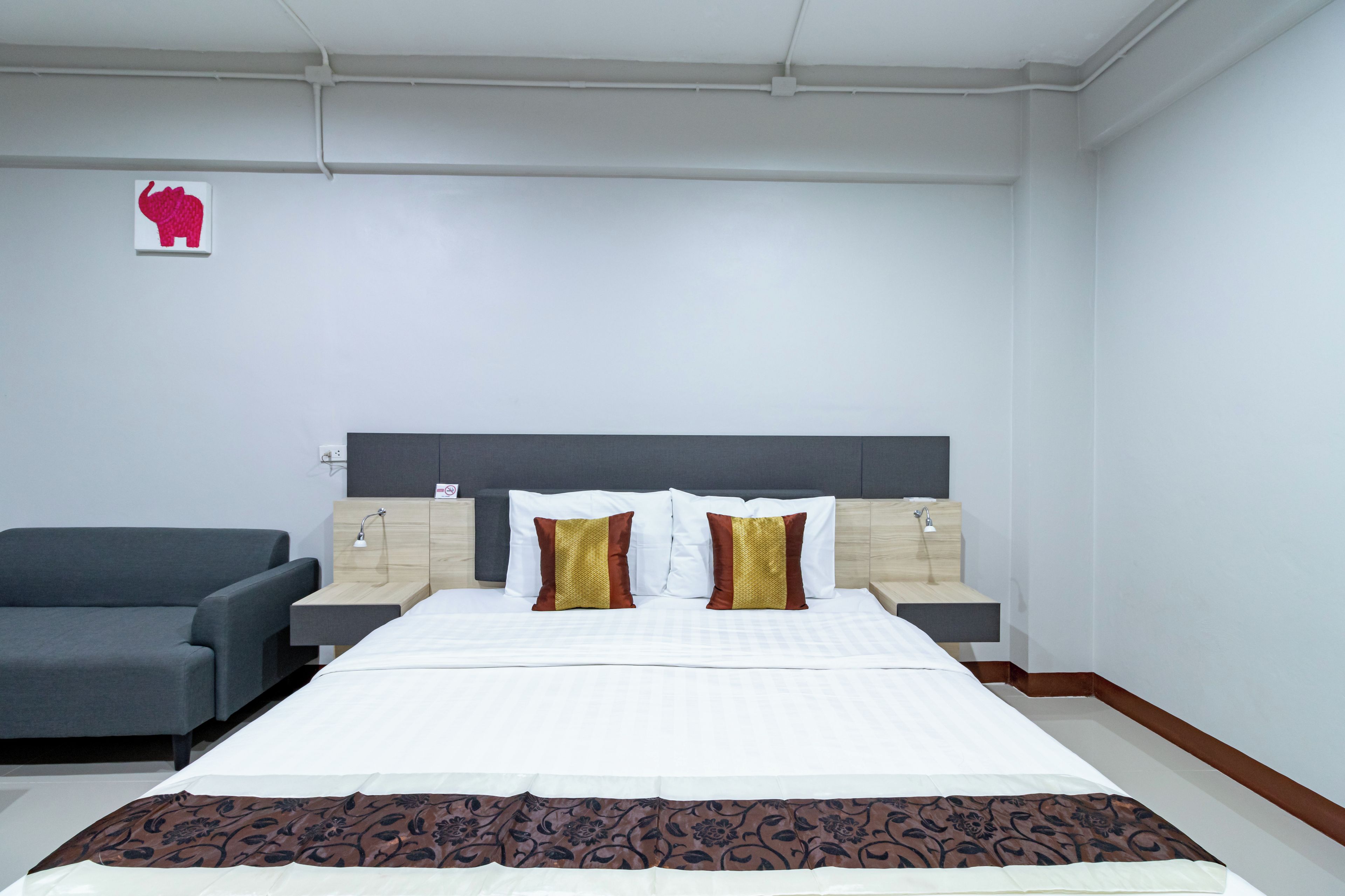 Habitación de hotel con una cama grande, dos mesitas de noche, un sofá gris y una decoración de elefante colgada en la pared.