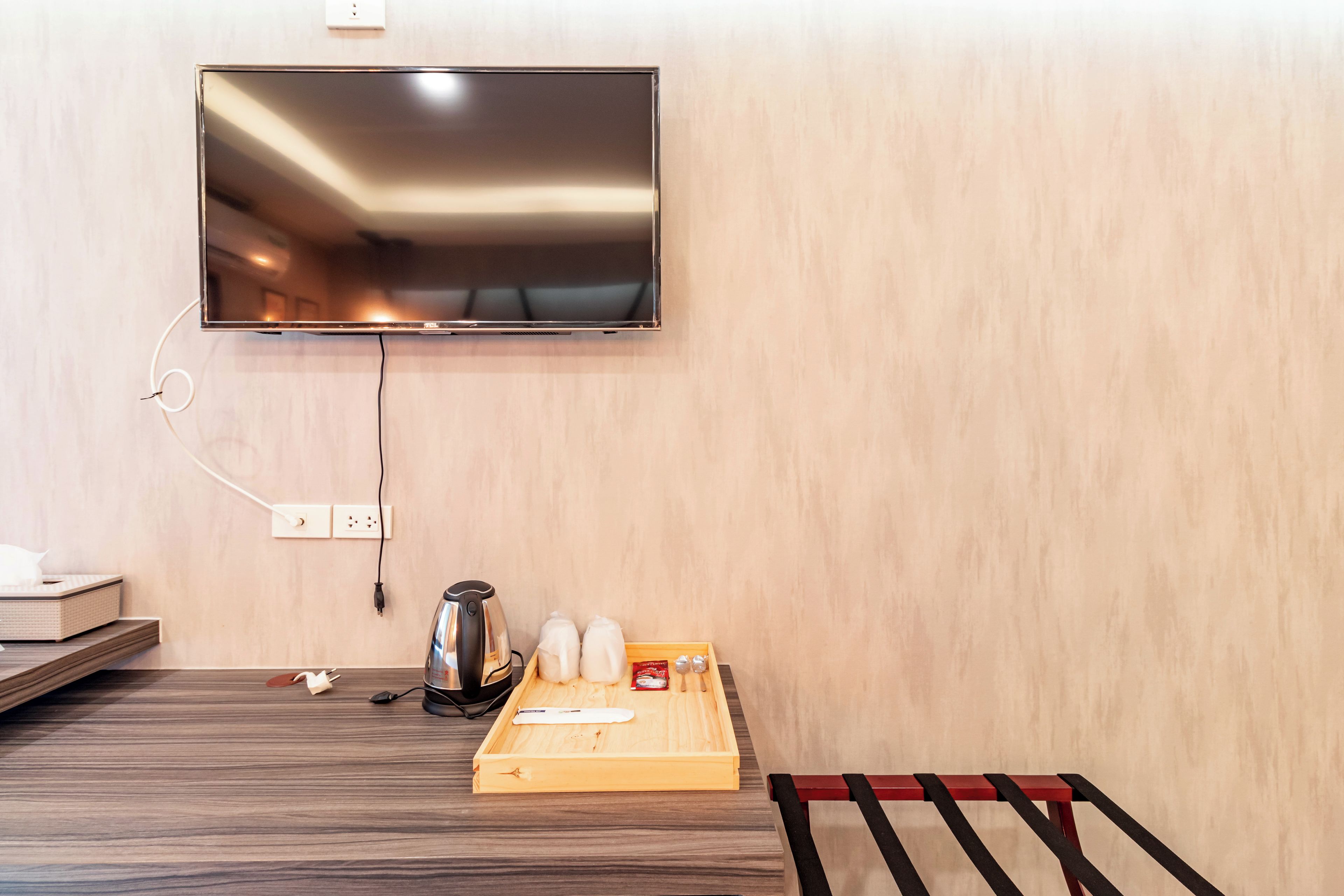 Habitación de hotel con televisor de pantalla plana en la pared, una bandeja de madera con infusiones y tetera, y un banco rojo.