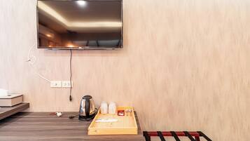 Habitación de hotel con televisor de pantalla plana en la pared, una bandeja de madera con infusiones y tetera, y un banco rojo.