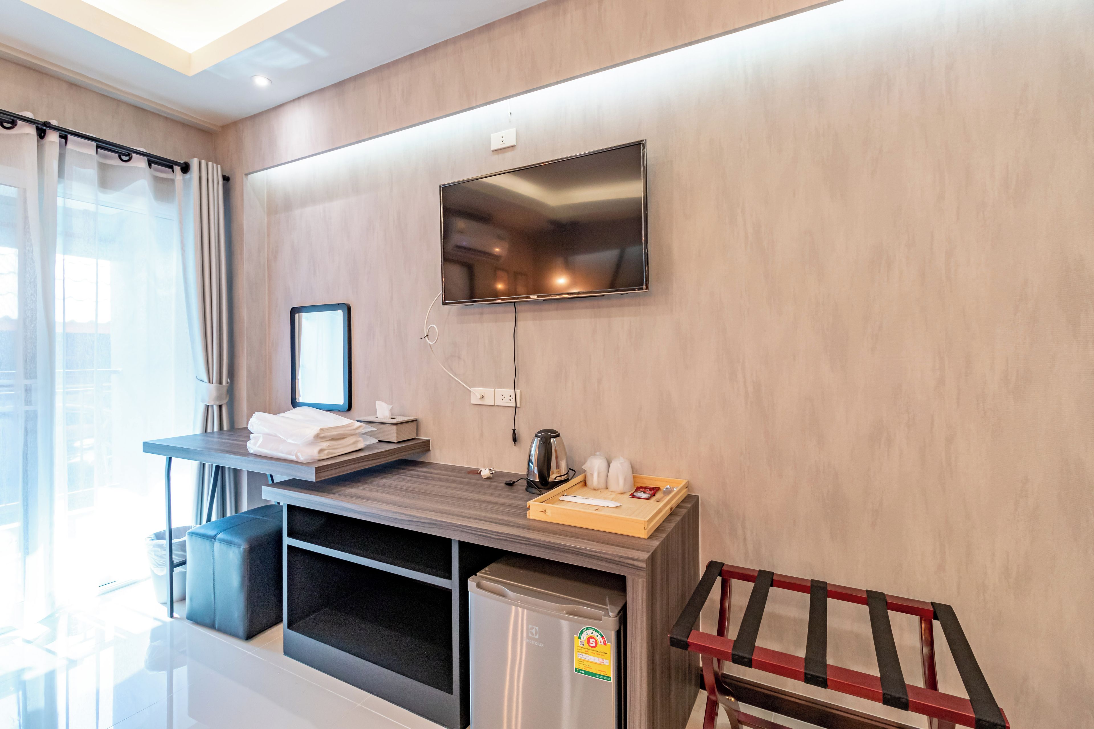 A modern hotel room with a flat-screen TV mounted on the wall, a small kitchenette with a refrigerator, and a seating area with a chair and a table.