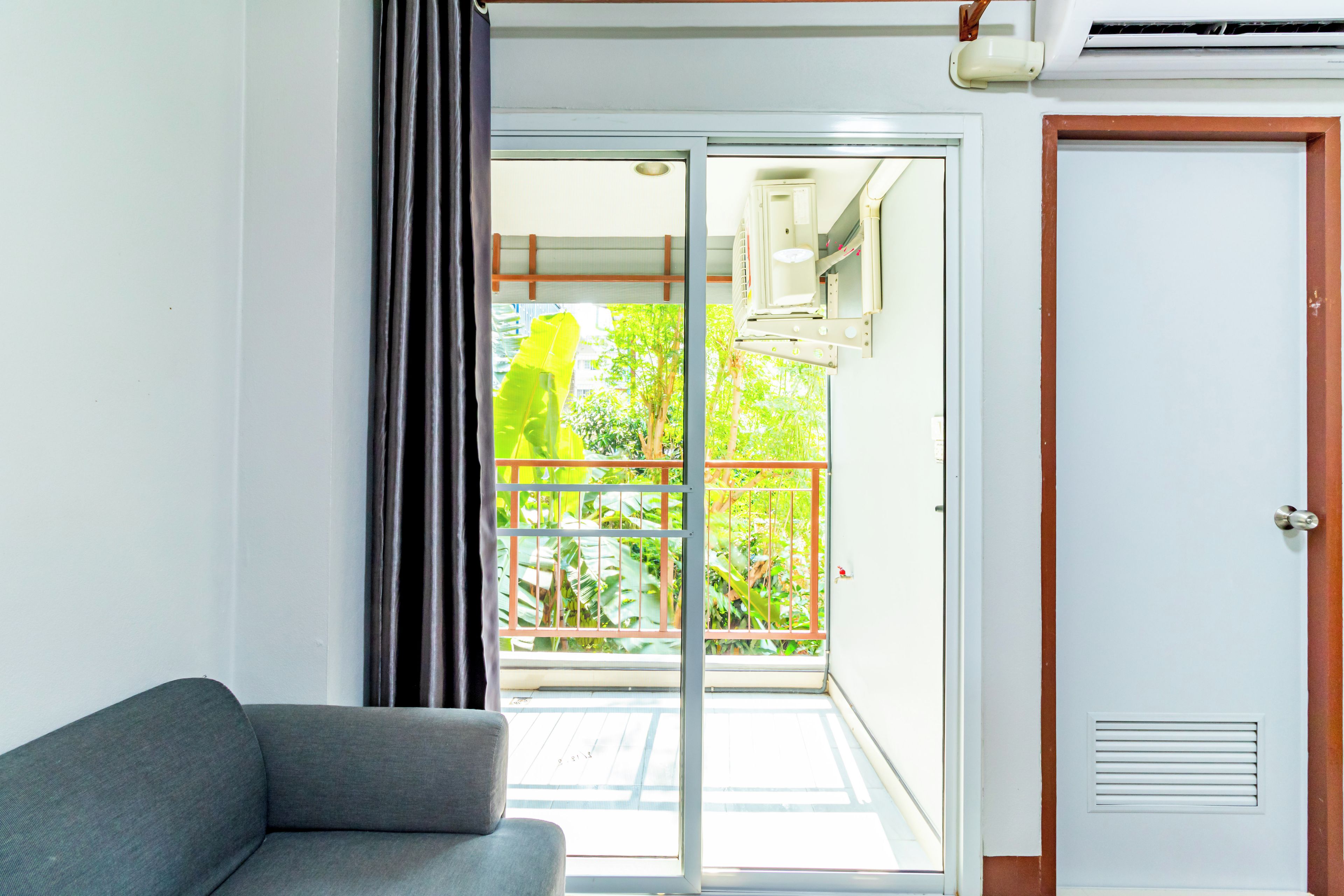 A room with a grey sofa, a sliding glass door leading to a balcony with a view of greenery, and a white door with a red trim.