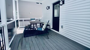 Een overdekt terras met een tafel en stoelen, een zwarte deur en een witte leuning.