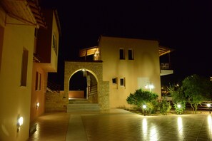 Área externa bem iluminada com um arco de pedra que dá acesso à entrada de um edifício, cercada por vegetação e iluminada por luzes nas paredes.