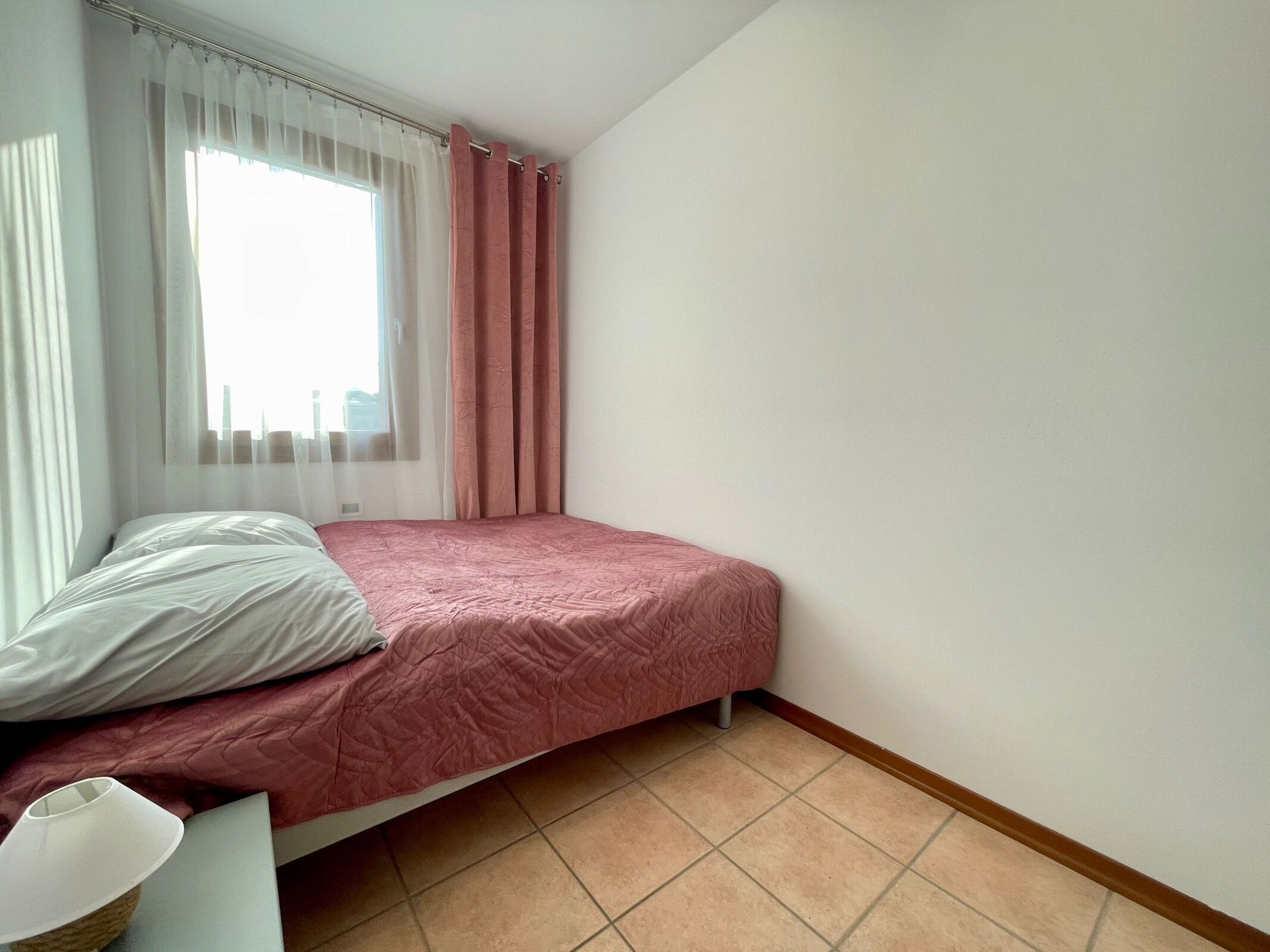 Un dormitorio con cama, mesita de noche, lámpara, ventana con cortinas y suelo de baldosas.