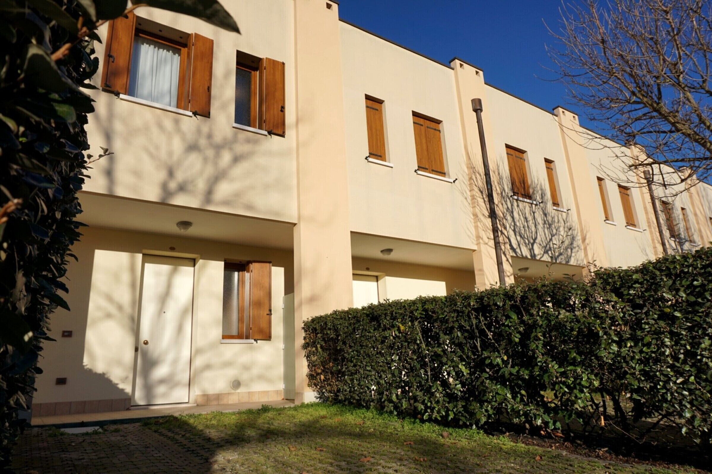 A row of modern, beige-colored apartment buildings with wooden shutters, a well-maintained hedge, and a paved driveway.
