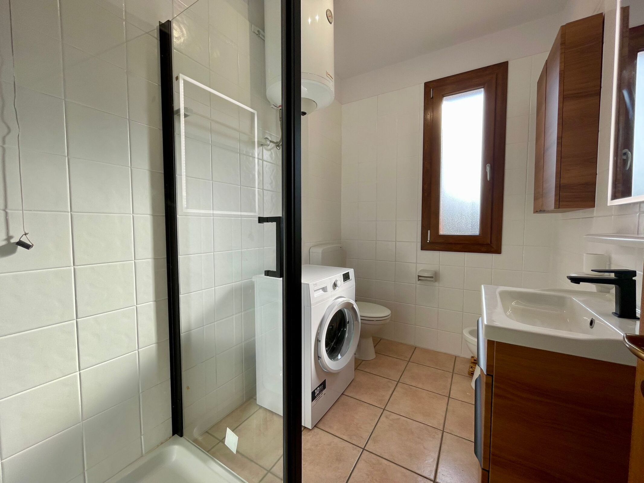 Et badeværelse med hvide fliser, en vaskemaskine, toilet og et håndvaskspejl.