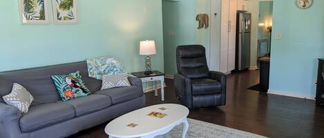 A living room with a gray sofa, a white coffee table, a black leather chair, and a kitchen in the background.