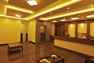A hotel lobby with a reception desk, a small seating area, and a staircase leading to an elevator.