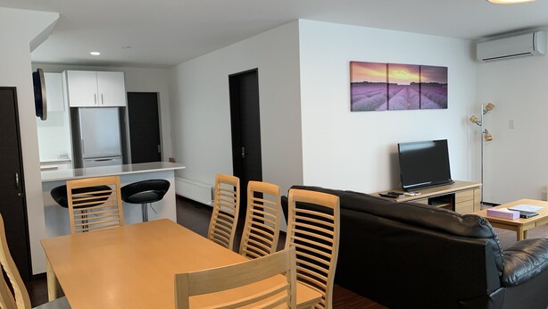A modern living room with a wooden dining table, four chairs, a black leather sofa, and a flat-screen TV. - Momiji Brooklyn (Furano)