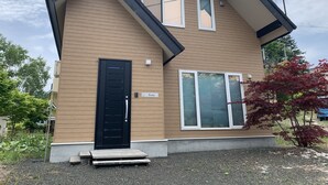 A modern two-story house with a black front door, a small porch, and a large window on the second floor. - Momiji Brooklyn (Furano)