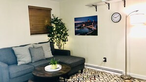 Un salon avec un canapé gris, une table basse ronde ornée d’une plante, un tapis à motifs, un tableau représentant un paysage urbain, une horloge et un lampadaire.