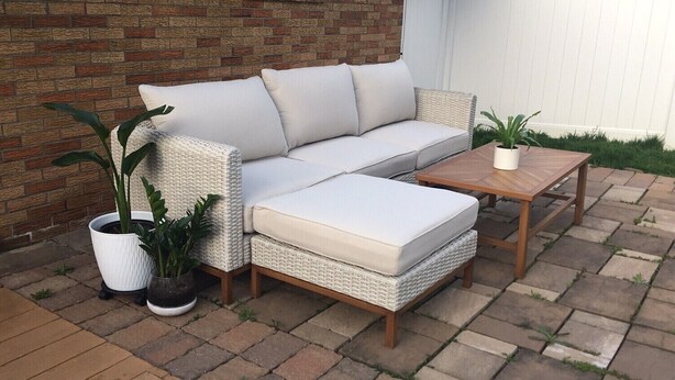 Un canapé en osier avec des coussins, une table basse en bois et des plantes en pot sur une terrasse, avec un mur de briques et une clôture blanche en arrière-plan.