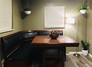 Un comedor con mesa de madera, banca acolchada negra y un cuenco de piñas. Hay dos plantas colgantes y una lámpara de pie.
