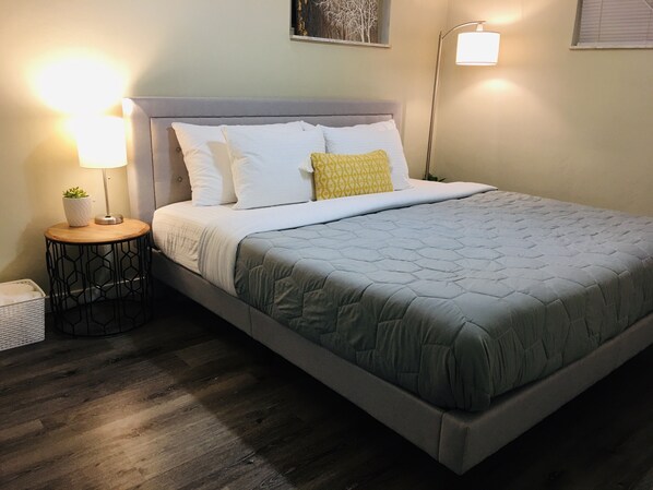 Una cama bien hecha con colcha gris, almohadas blancas y una almohada de color amarillo. Hay una mesita de noche con una lámpara y una pequeña planta en maceta. La habitación tiene suelo de madera y pared de color claro.