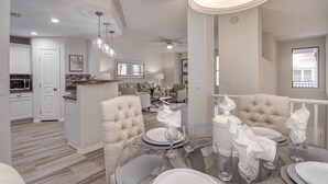 A modern dining area with a glass table set for four, surrounded by plush armchairs. A kitchen with a microwave and a dining island with a sink is visible in the background.