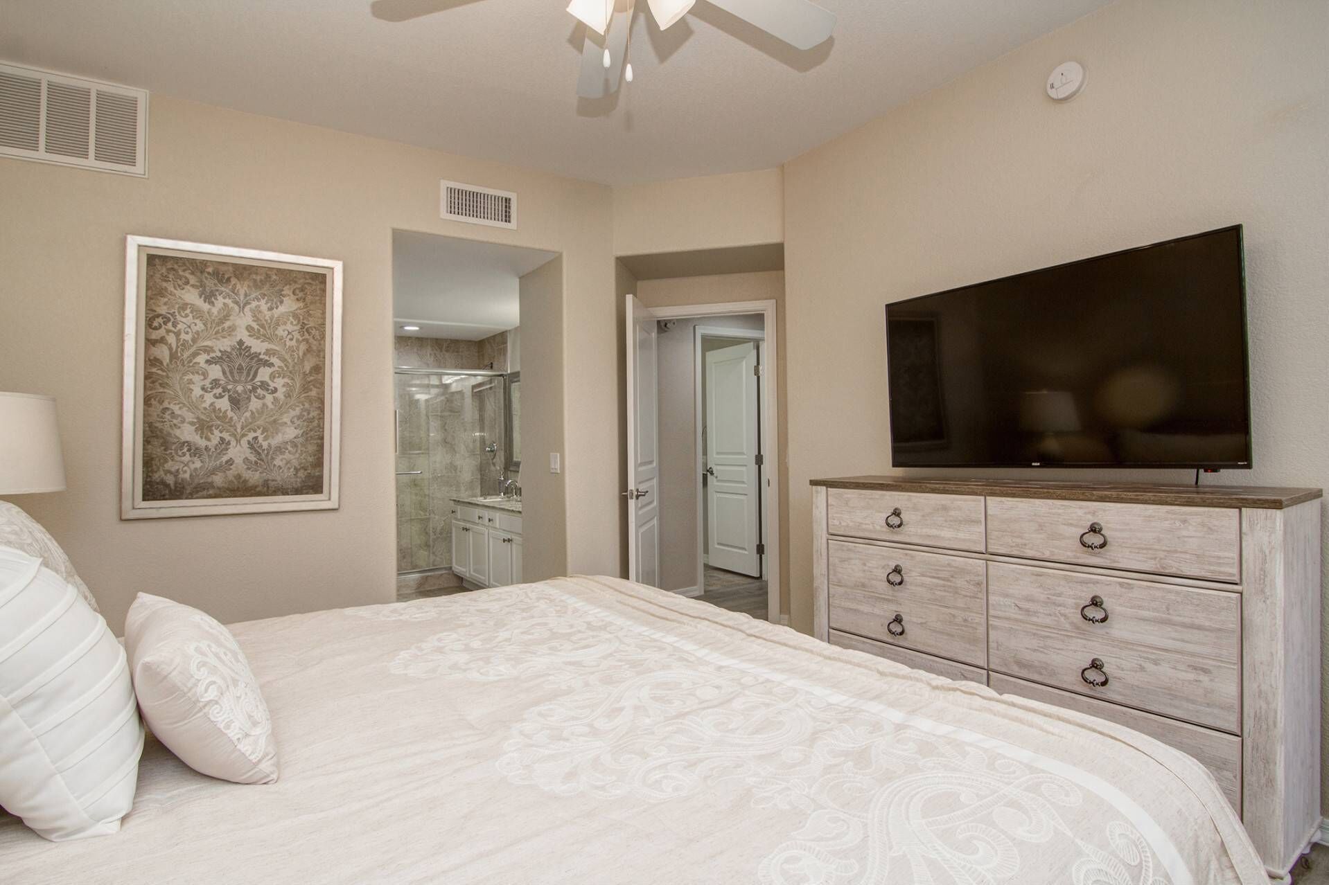 A bedroom with a bed, a TV, a dresser, a shower, and a door leading to another room.