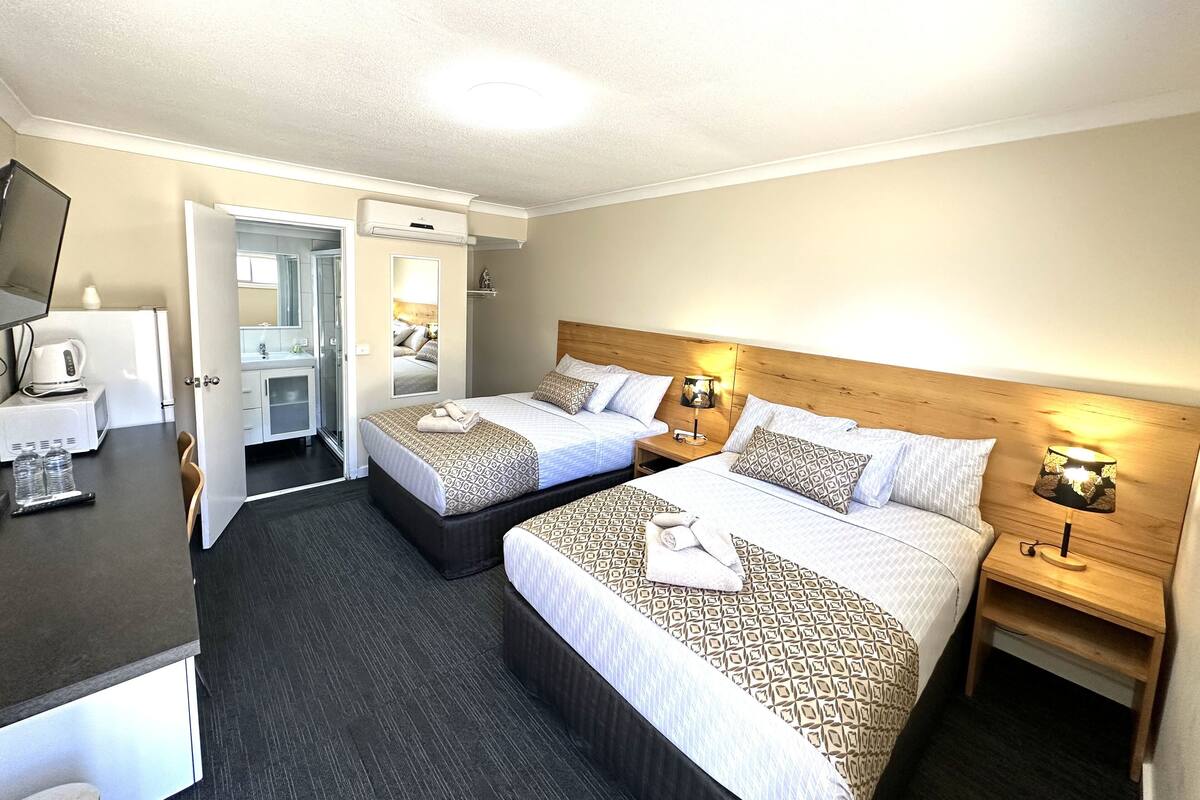 Quarto de hotel com duas camas, uma escrivaninha, um espelho e uma pequena cozinha.