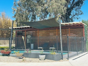A pool area with a covered seating area, a fenced pool, and surrounding plants.