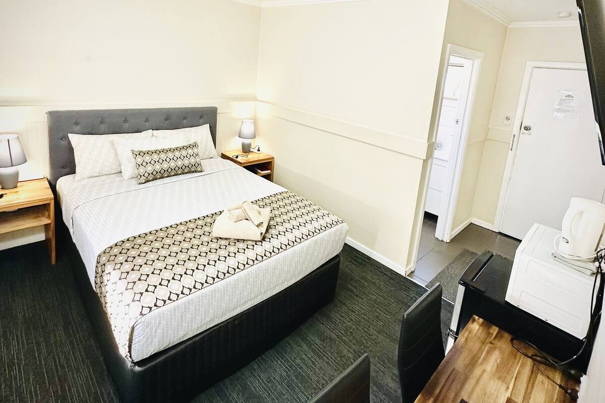 Quarto de hotel com uma cama grande, mesas de cabeceira com abajures, uma escrivaninha com computador e uma geladeira.