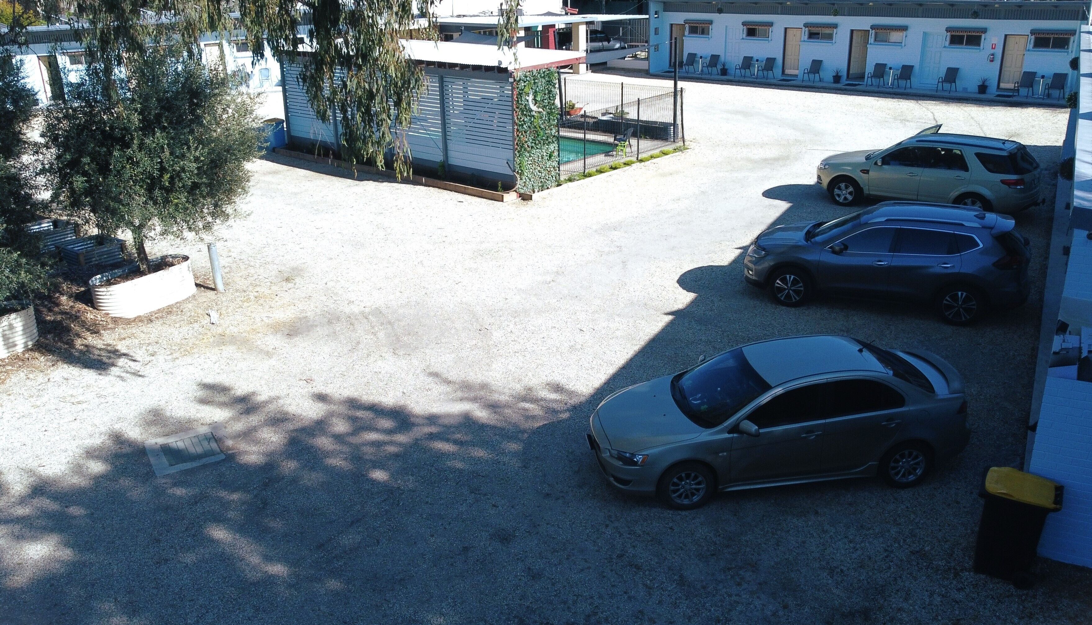 Un estacionamiento con tres coches, un pequeño jardín cercado y un edificio con múltiples ventanas y puertas.