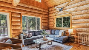 A log cabin interior with a sofa, armchair, and coffee table, large windows, and a ceiling fan. - Stargazer Lodge | Hot Tub + Game Loft (Glacier)