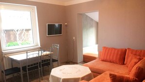 Um quarto com uma mesa de jantar e cadeiras, uma pequena mesa redonda com toalha de renda, um sofá laranja e uma TV fixada na parede.