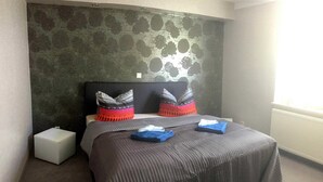 Cama bem arrumada com colcha cinza e duas almofadas, uma pequena mesa de cabeceira e papel de parede estampado atrás da cama.