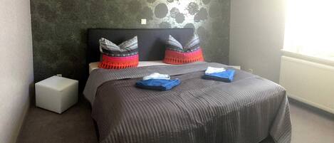 Una cama bien hecha con una colcha gris y dos almohadas, una mesita de noche pequeña y un papel tapiz estampado detrás de la cama.