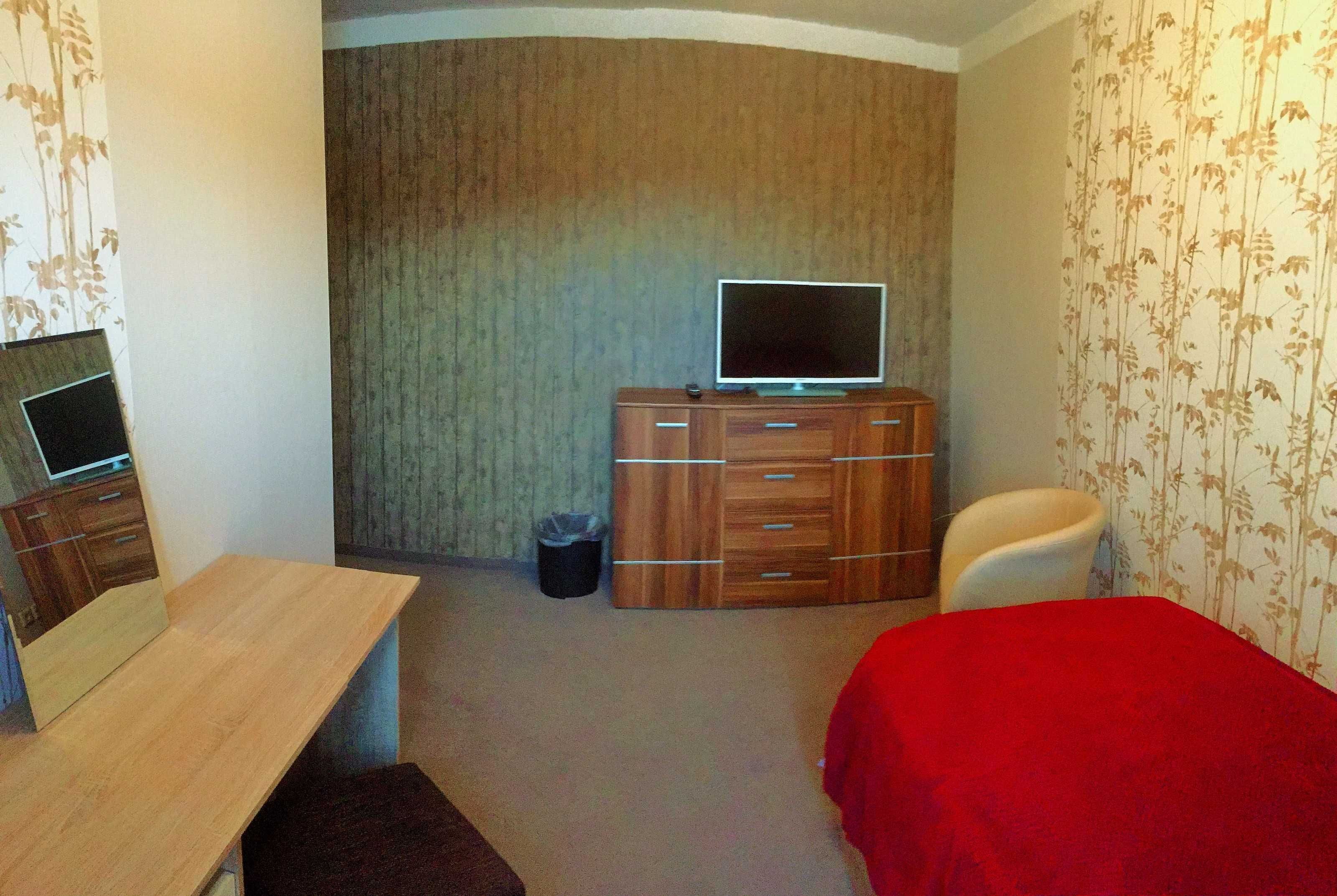 A bedroom with a wooden dresser, a television on top, a red bedspread, a chair, and a small table.