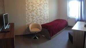 Una stanza piccola e arredata in modo essenziale, con un letto singolo, una sedia, una scrivania e una finestra.