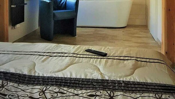 Um quarto pequeno com telhado inclinado, uma cama, uma cadeira azul, uma banheira e uma janela com cortinas.