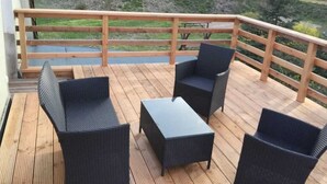Um deck de madeira com móveis de vime e guarda-corpo, com vista para uma área gramada onde estão estacionados veículos.