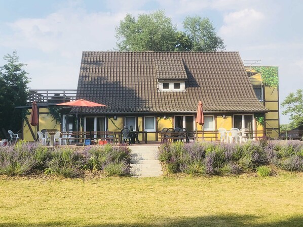 Un bâtiment à deux étages, doté d’un toit en tuiles, d’un espace extérieur aménagé avec des chaises et des tables, et d’un jardin où poussent des plants de lavande.