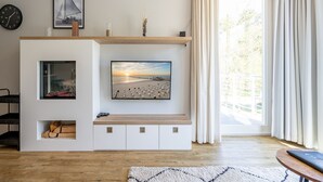 Un soggiorno con una grande televisione a schermo piatto montata su una parete bianca, una panca in legno, un camino e un'ampia finestra con tende.