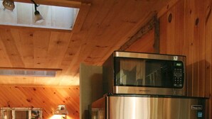 A compact kitchen with a stainless steel refrigerator and microwave, wooden walls, a dining table with chairs, and a small rug. - Adirondack Riverfront Retreat (Jay)