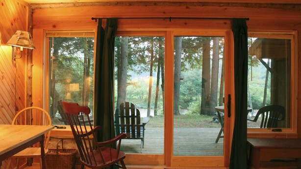 A wooden cabin with a sliding glass door leading to an outdoor deck. Inside, there is a wooden table and two rocking chairs. - Adirondack Riverfront Retreat (Jay)