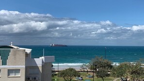 A coastal view with a large cargo ship in the distance, a building with a flat roof, and a clear sky.
