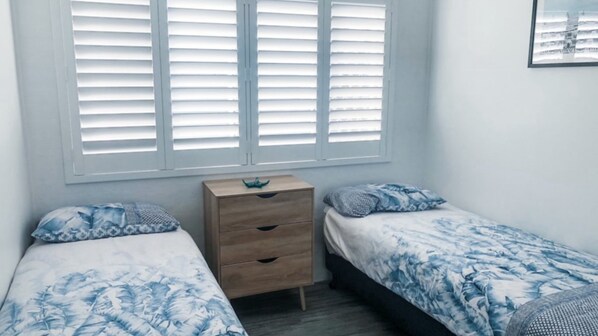 A bedroom with two single beds, a wooden dresser, and a window with blinds.