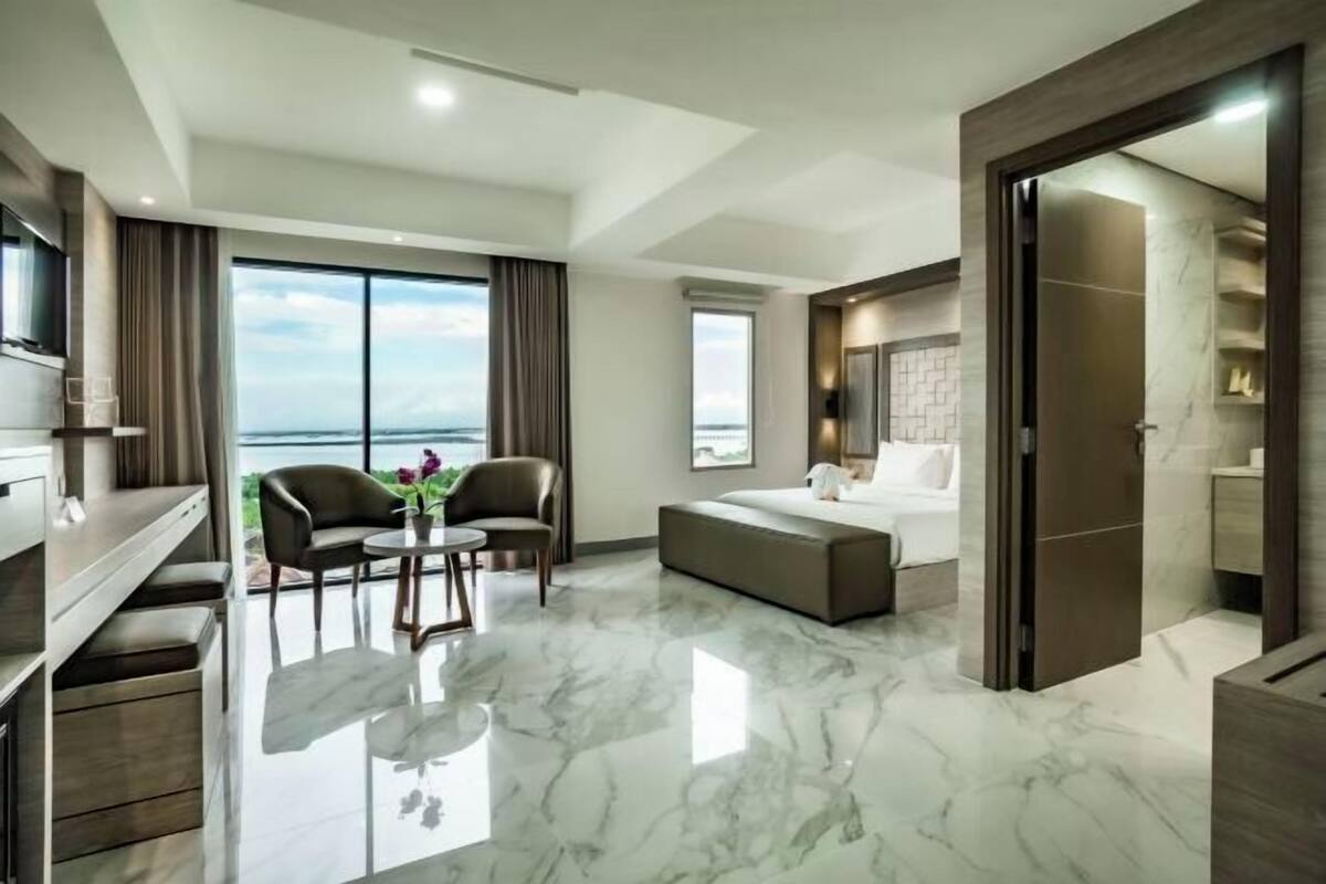 A modern hotel room with a large window, a bed, a seating area with a table, and a TV.