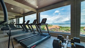 Une salle de sport équipée de tapis de course et d’haltères, avec de grandes fenêtres offrant une vue sur la mer et les bâtiments.