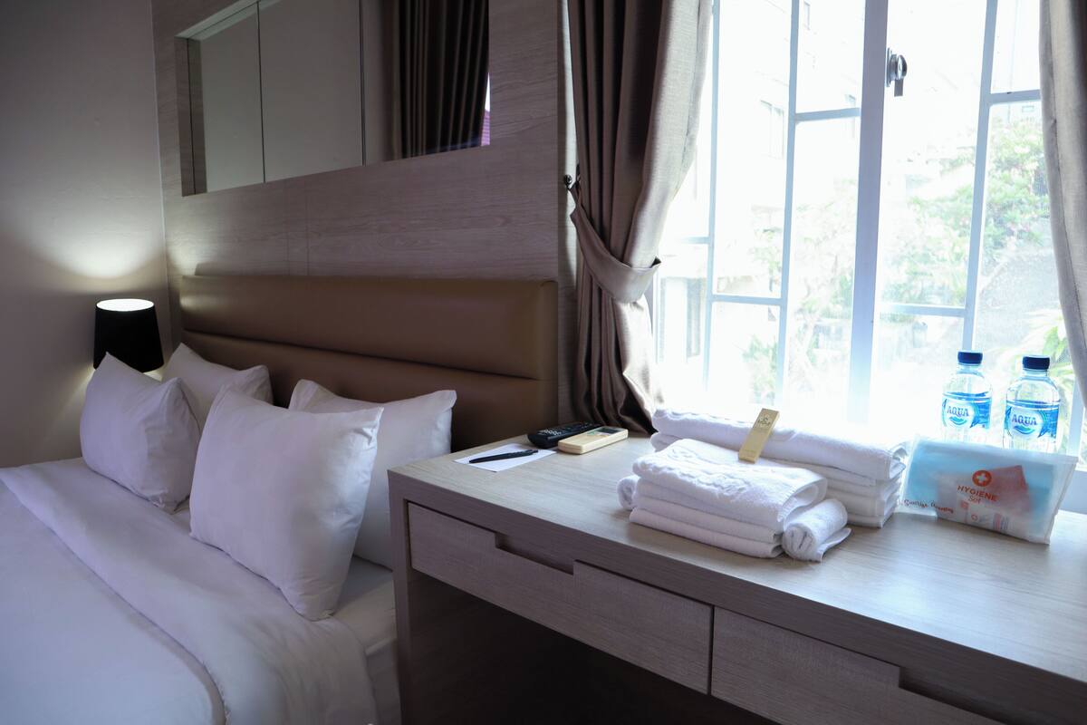 A hotel room with a bed, a bedside table with toiletries, a nightstand, and a window with curtains.