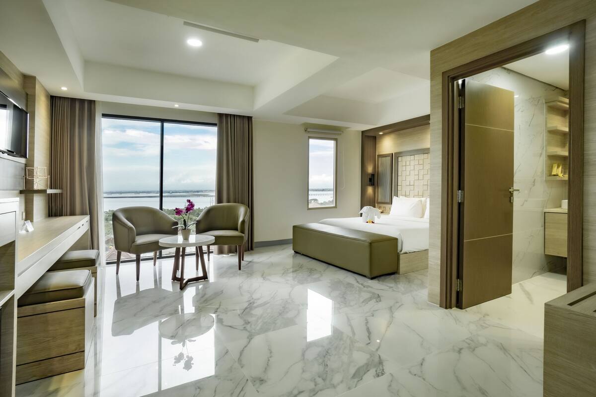 A modern hotel room with a large bed, a seating area with two chairs, a small table with flowers, and a view of the sea through large windows.