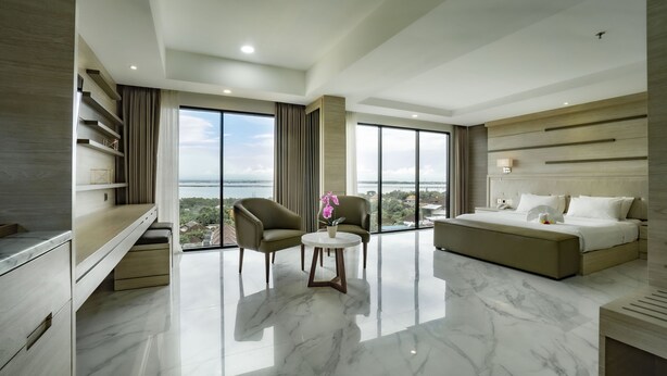 Une chambre d’hôtel moderne avec un grand lit, un coin salon comprenant deux fauteuils et une petite table, et une vue sur la ville grâce à de grandes fenêtres.