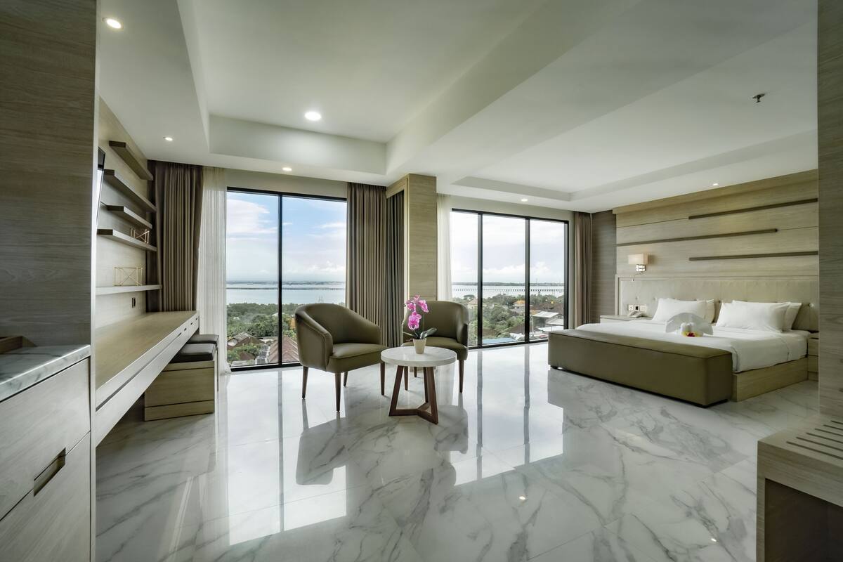 A modern hotel room with a large bed, a seating area with two chairs, a small table, and a view of the city through large windows.