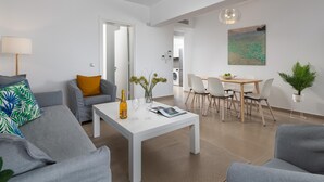 Ein modernes Wohnzimmer mit einer Couch, einem Couchtisch mit Flasche und Gläsern, einem Essbereich mit Stühlen und Tisch sowie einer Küche im Hintergrund.